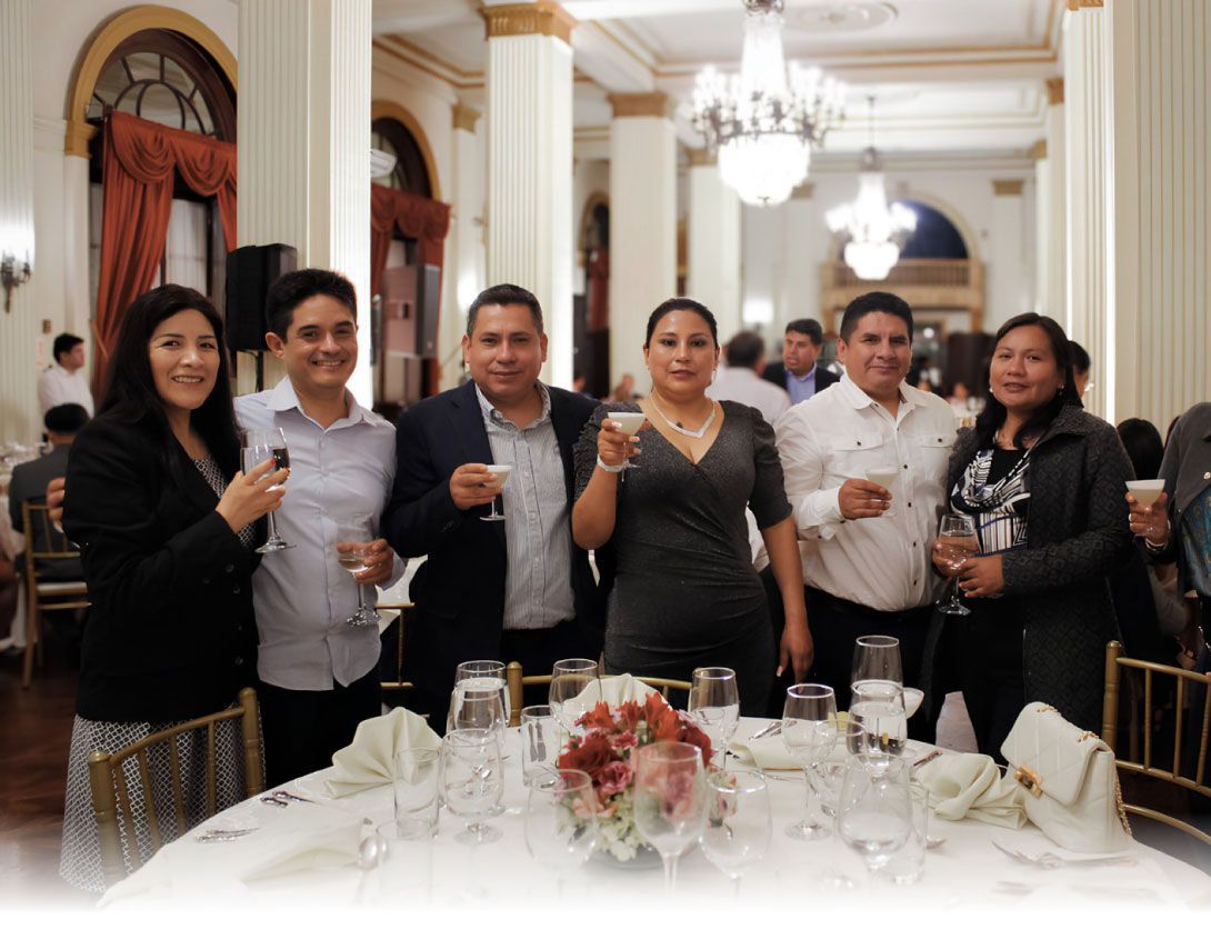 Grupo de seis personas brindando con bebidas en un evento formal; mesa preparada con flores y lámpara de araña.