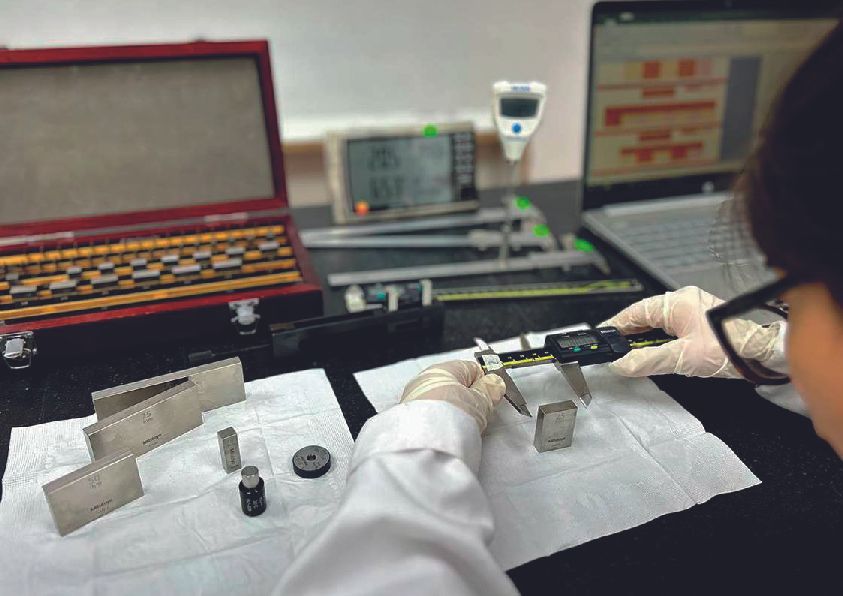 Persona con bata de laboratorio que utiliza calibradores digitales en piezas metálicas en una estación de trabajo.