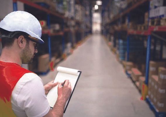 Trabajador de almacén con casco, escribiendo en el portapapeles, comprobando el inventario.