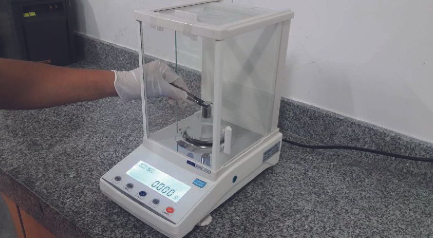 Una mano enguantada que utiliza pinzas para colocar un elemento en una balanza de laboratorio.