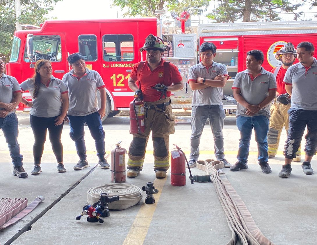 Bomberos frente a camiones de bomberos, uno de ellos con uniforme rojo muestra el equipo.