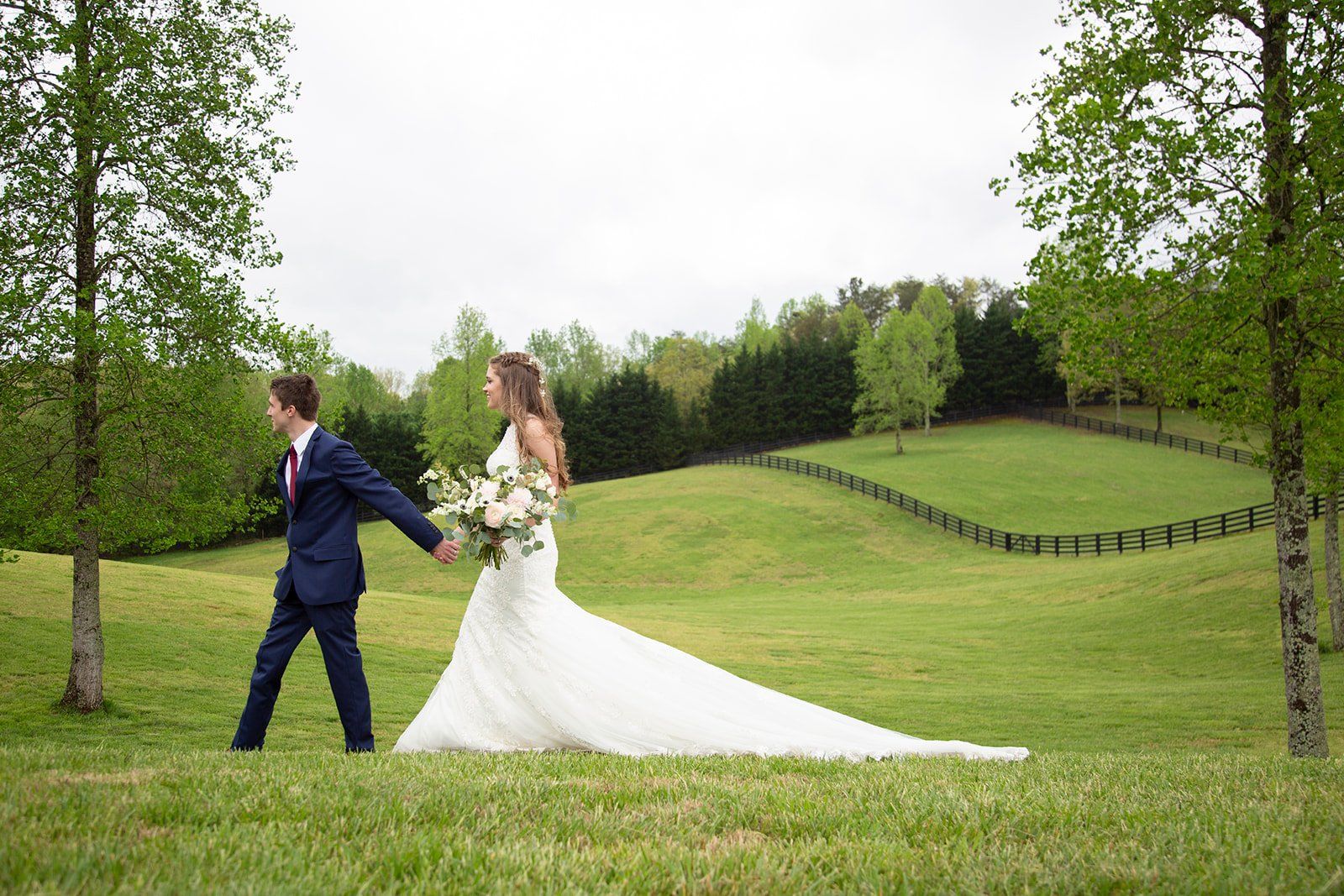 A bride and groom are walking through a grassy field holding hands.