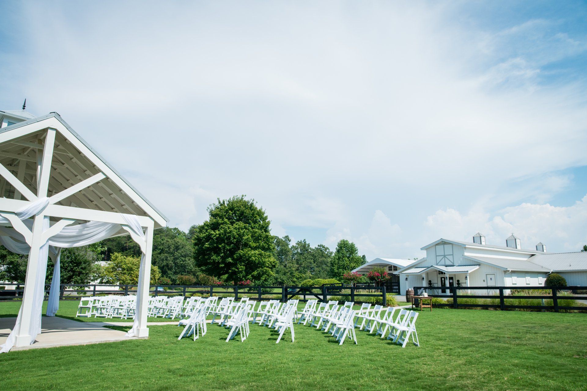 A row of white folding chairs are lined up in a grassy field in front of a white gazebo.