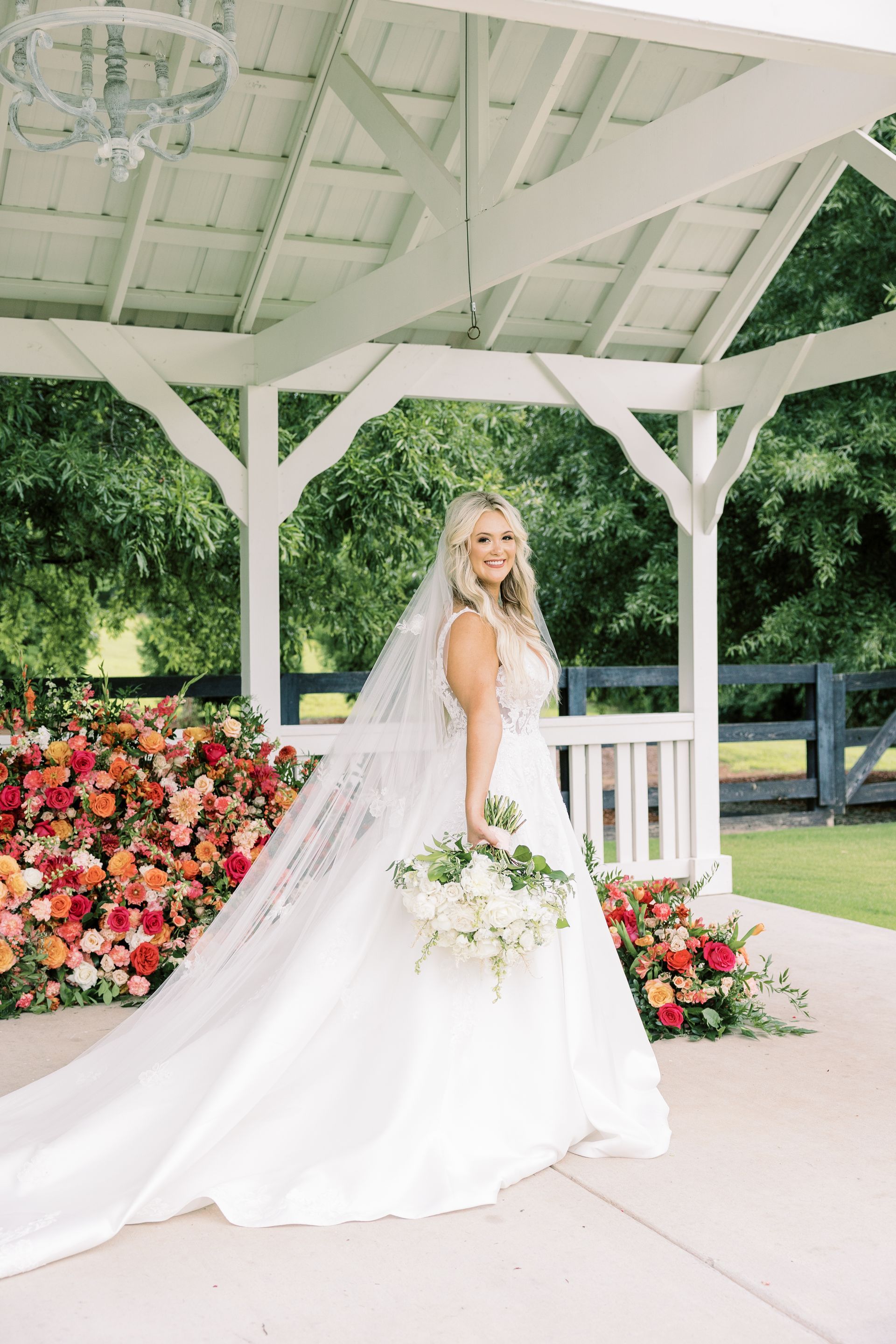 A bride in a wedding dress and veil is standing under a gazebo holding a bouquet of flowers.