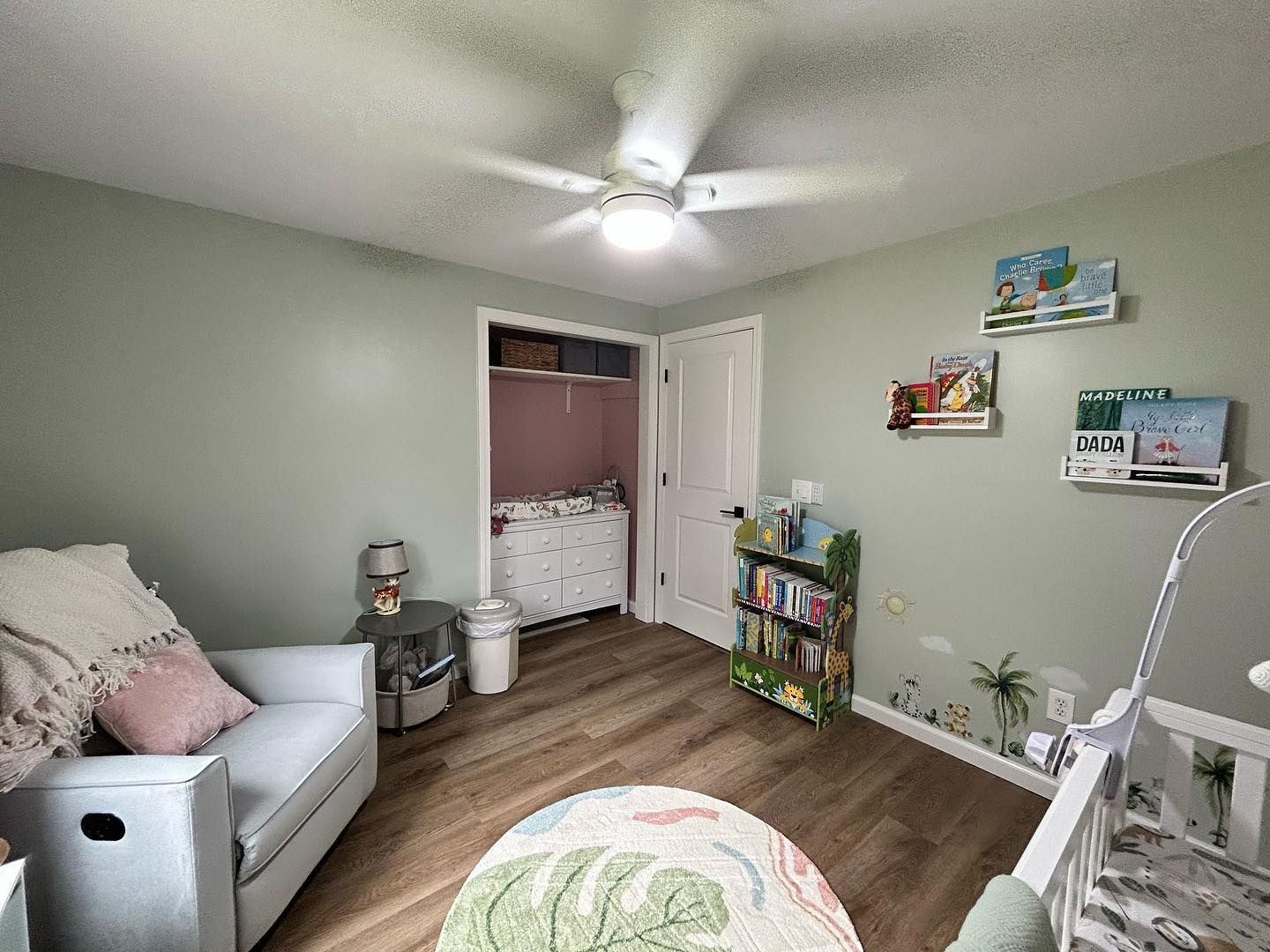 A nursery with light blue walls, a glider, crib, and bookshelf. The room has light wood flooring.