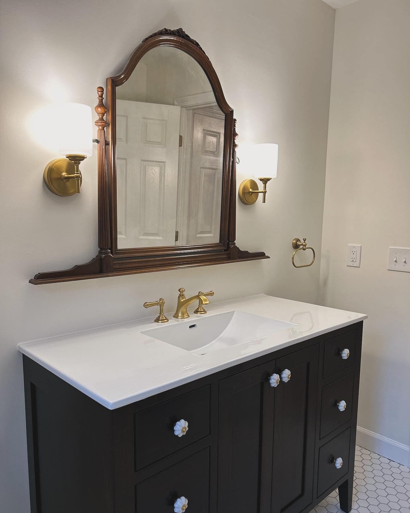 Bathroom vanity with dark brown cabinet, white countertop, gold fixtures, and arched mirror.