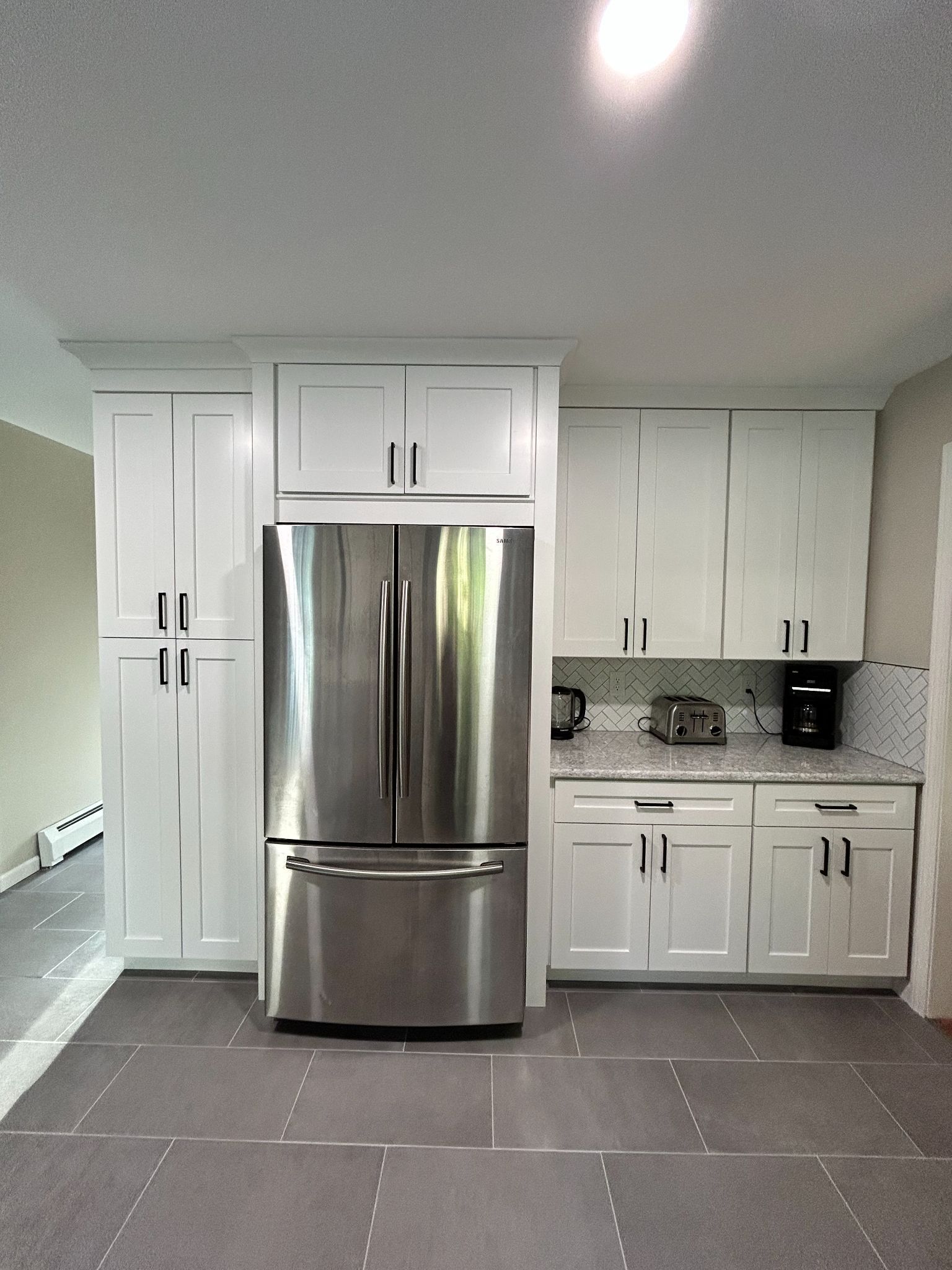 White kitchen cabinets surround a stainless steel refrigerator. Gray countertops and flooring.