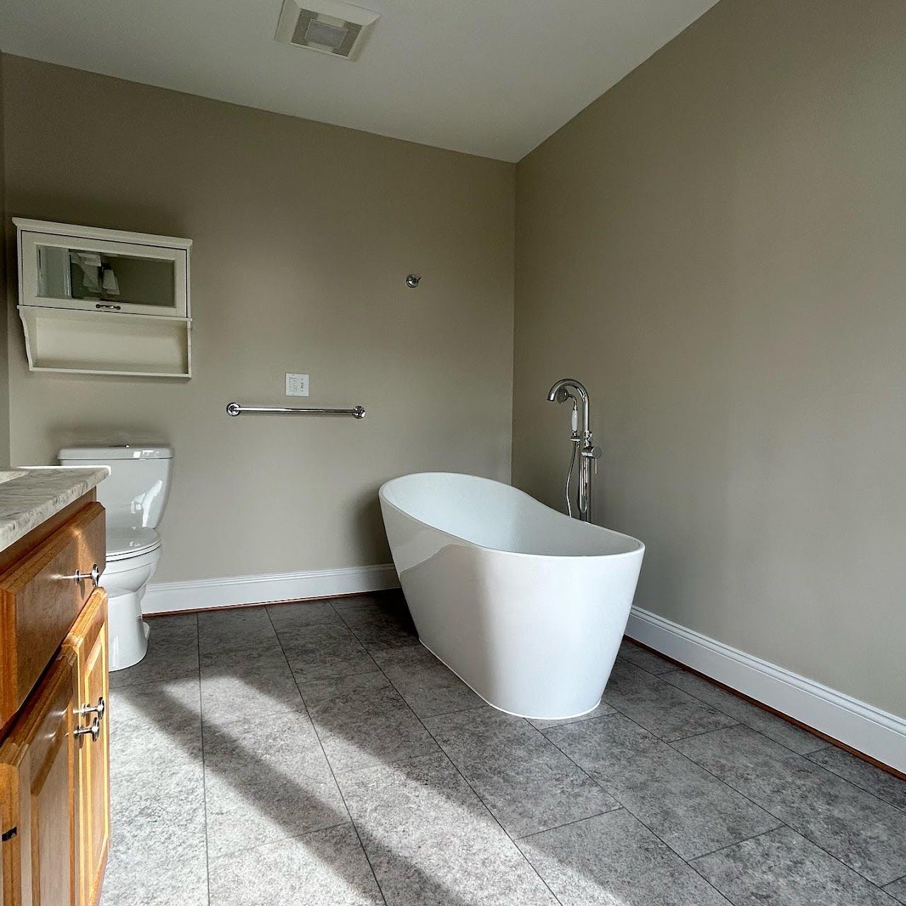 Bathroom with a white freestanding tub, toilet, and vanity. Gray tile floor, light tan walls, and a silver towel bar.