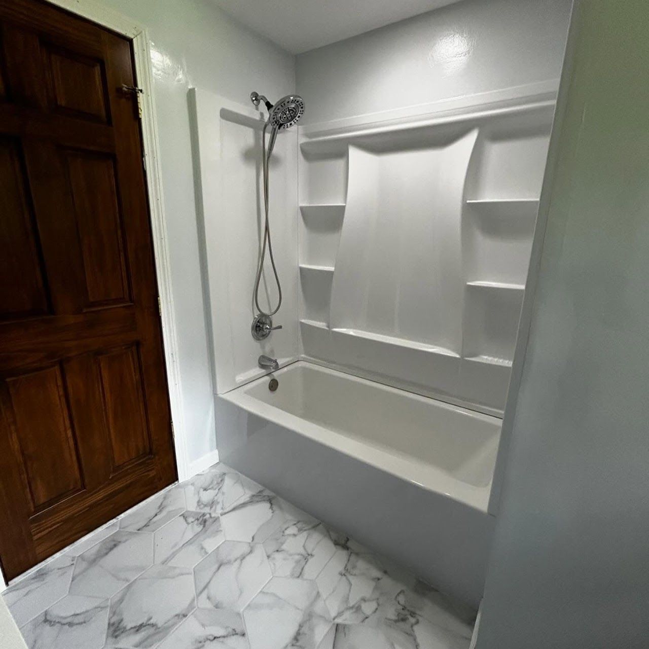 White bathroom with a tub, shower, and marble-patterned floor next to a dark wooden door.