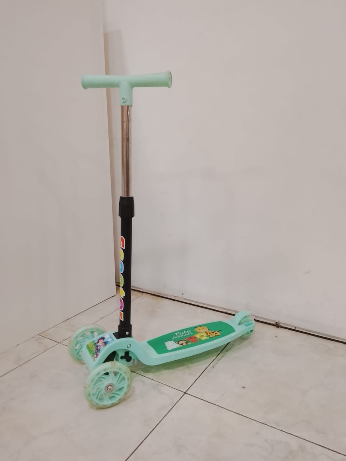 Un patinete de tres ruedas de color verde claro está arrinconado contra una pared y un suelo blancos.