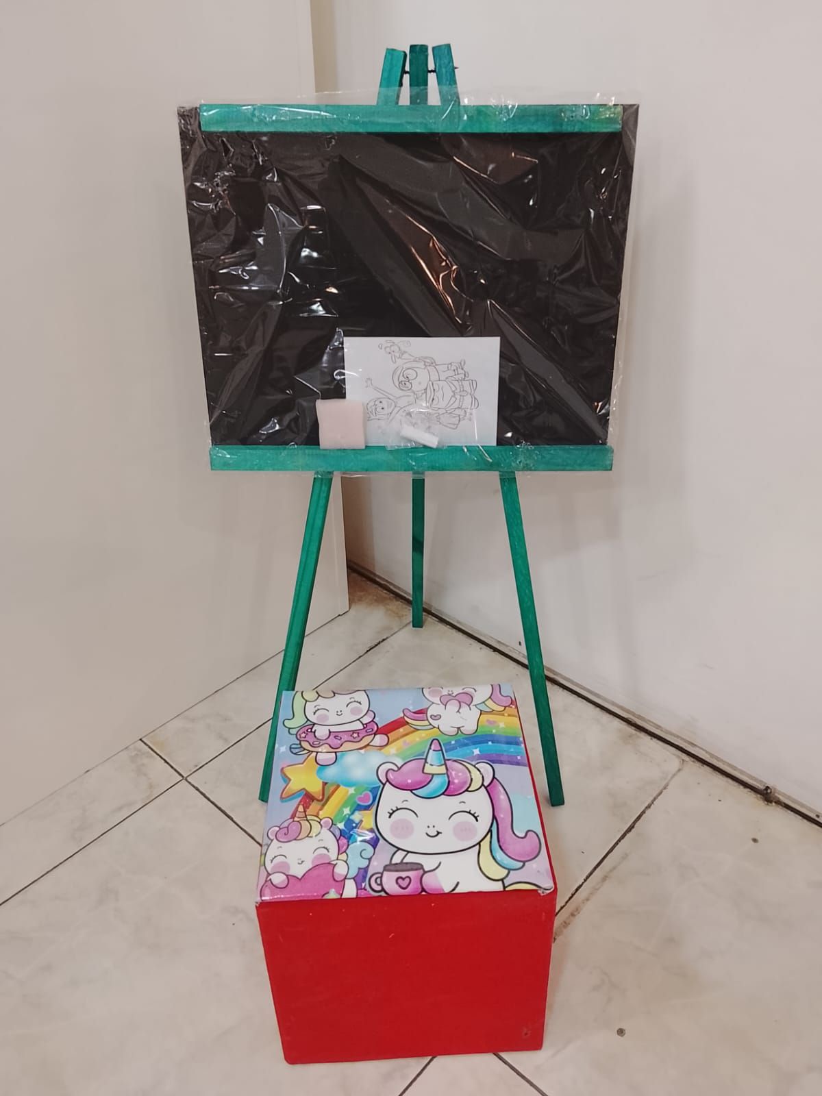 Caballete verde con superficie negra, una página para colorear y una caja de almacenamiento roja con temática de unicornio.