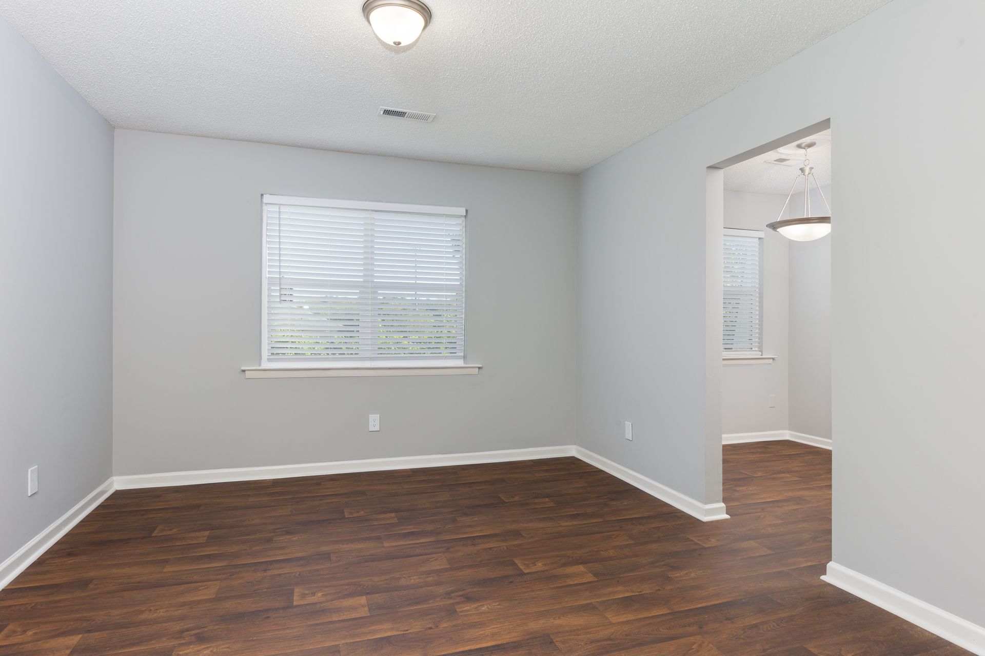 Empty room with light gray walls, dark wood-look floors, a white window with blinds, and a doorway to another room.
