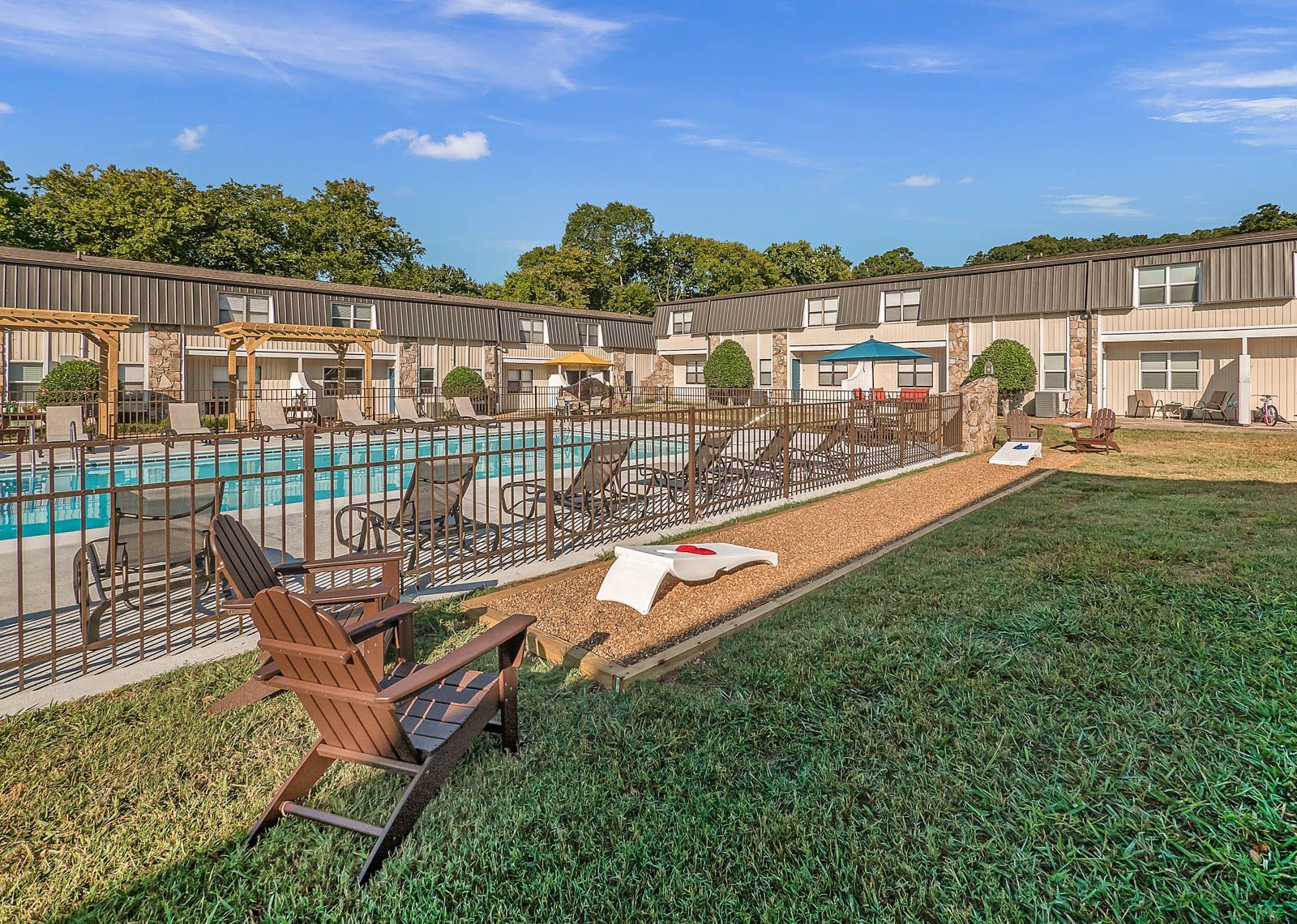 A swimming pool area surrounded by lounge chairs, wooden pergolas, and low-rise residential buildings under a blue sky.