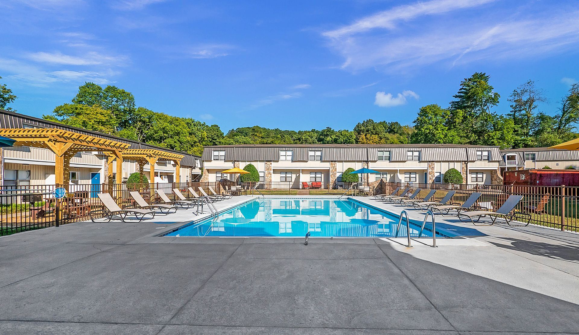 An outdoor swimming pool surrounded by lounge chairs, a wooden pergola, and apartment buildings under a clear blue sky.