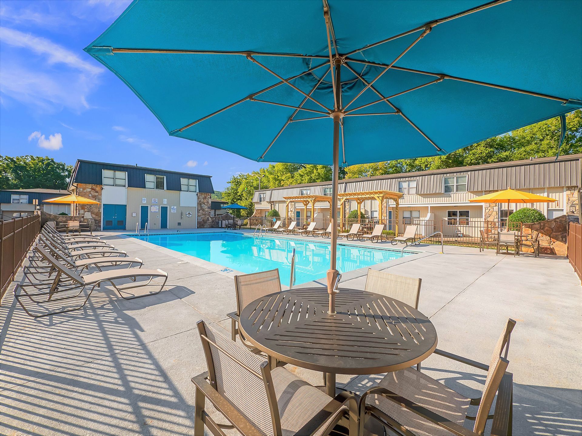 A sunny outdoor swimming pool area with lounge chairs, dining tables under umbrellas, and residential buildings.