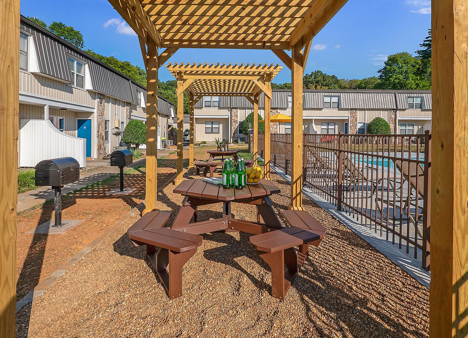 Outdoor picnic area with wood tables and grills under a pergola, adjacent to an apartment complex and pool.
