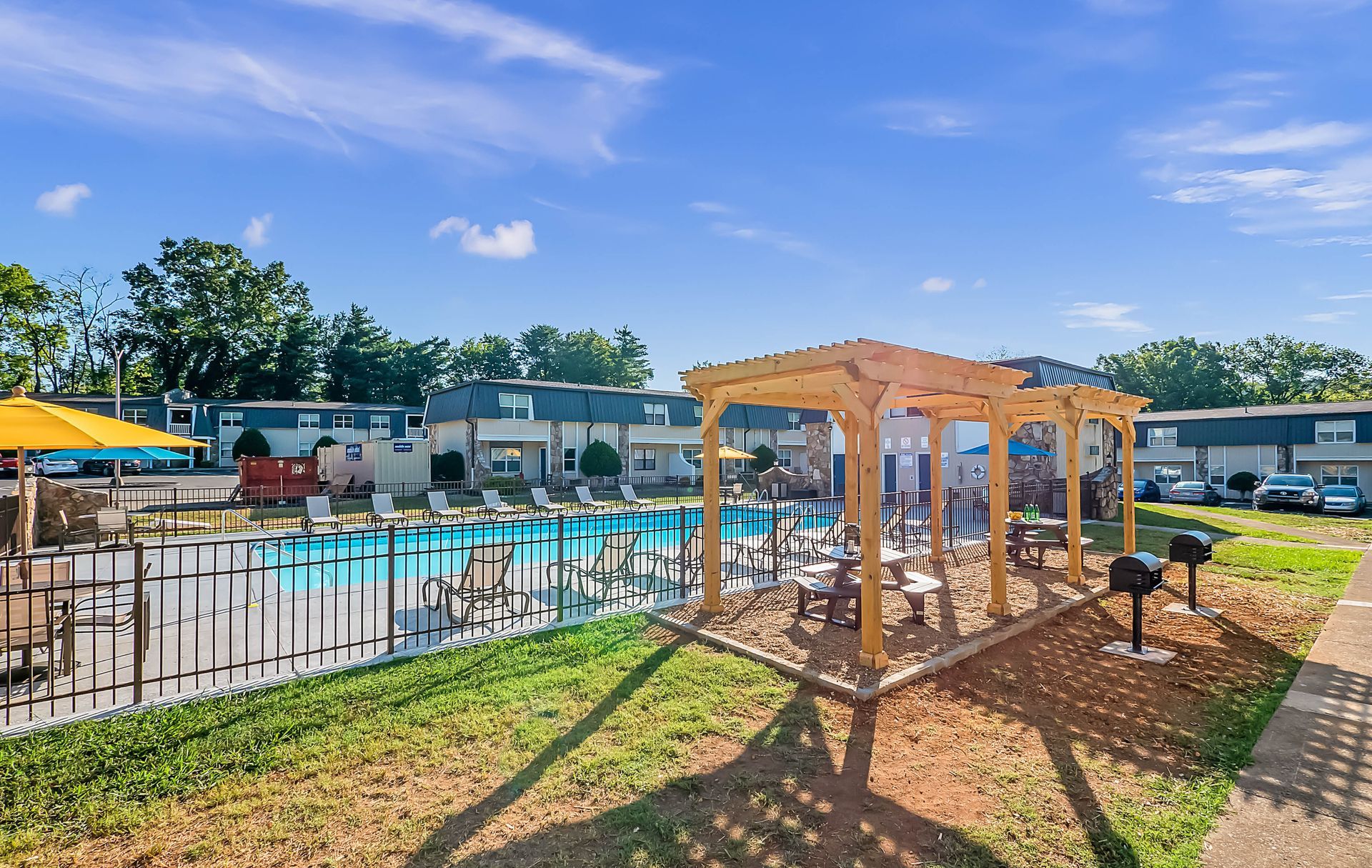 An outdoor pool area with a wooden pergola, barbecue grills, and apartment buildings under a clear blue sky.