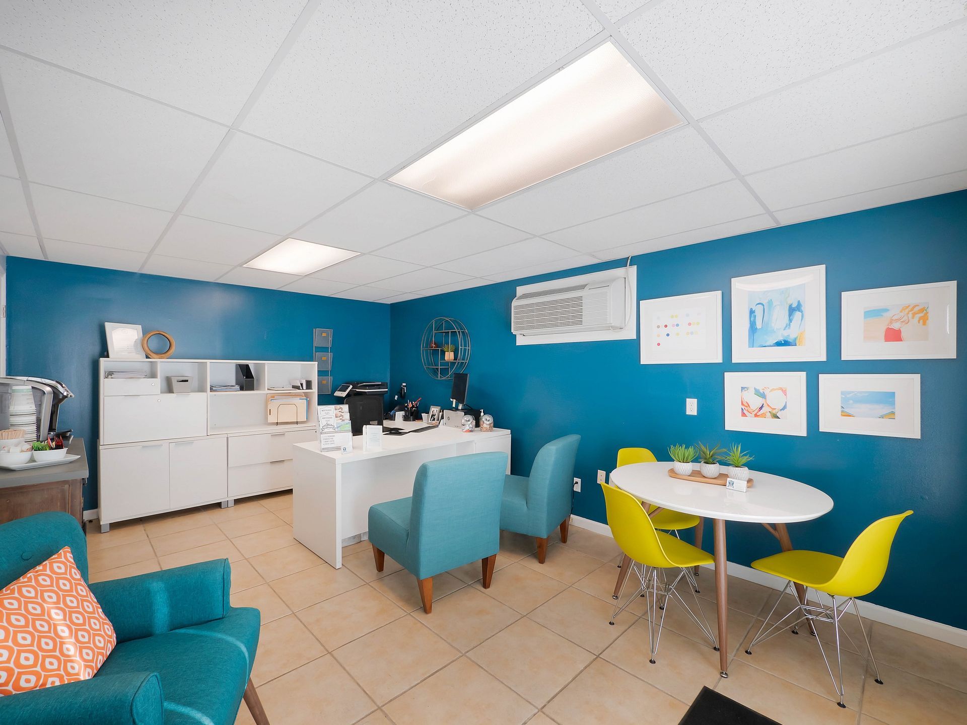 A bright office with teal walls, a white desk, two teal chairs, a round table with three yellow chairs, and wall art.