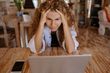 Woman with curly hair looks stressed, hands on head, staring at a laptop on a wooden table.