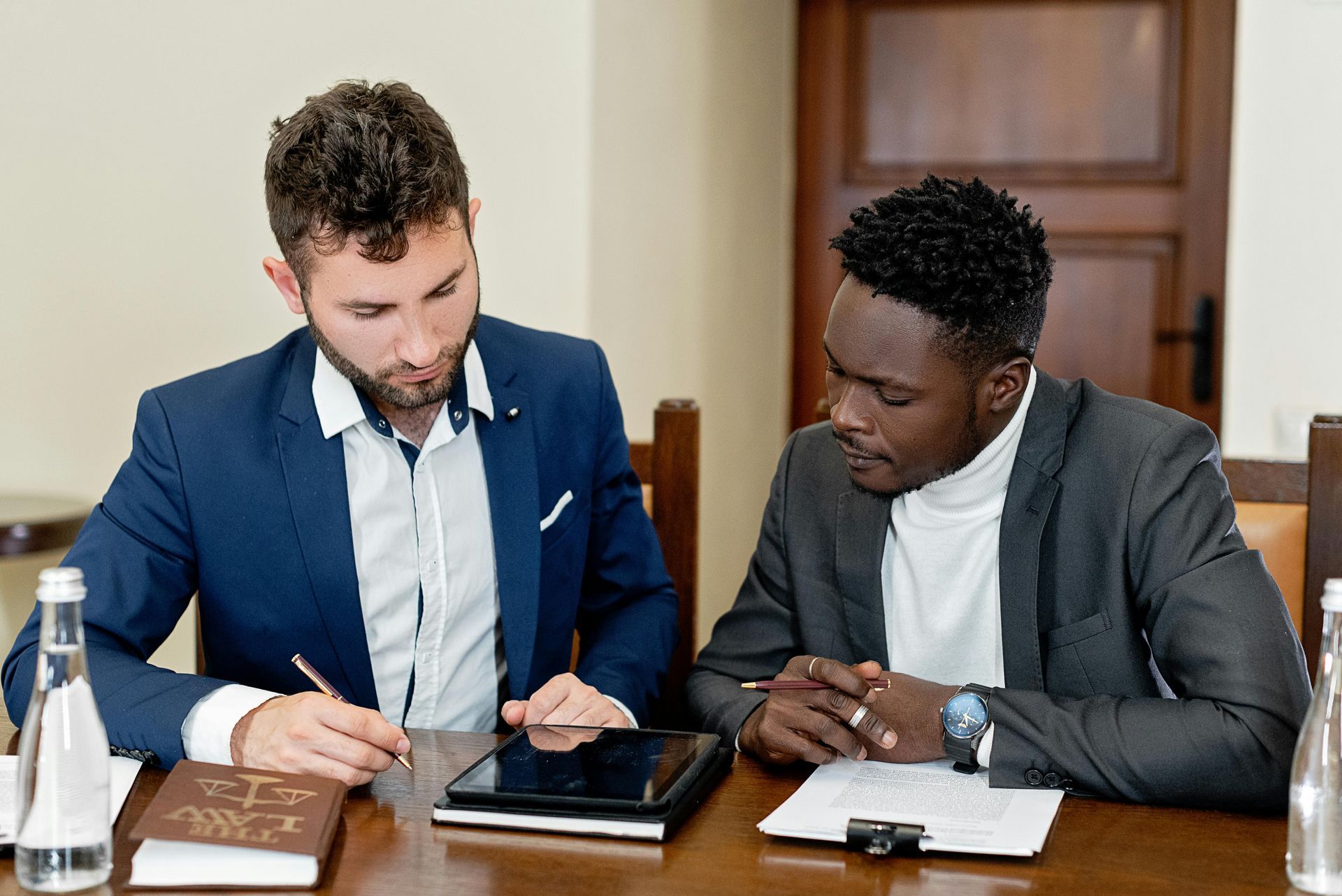 Two men in suits review documents at a table. One signs a paper, the other watches.