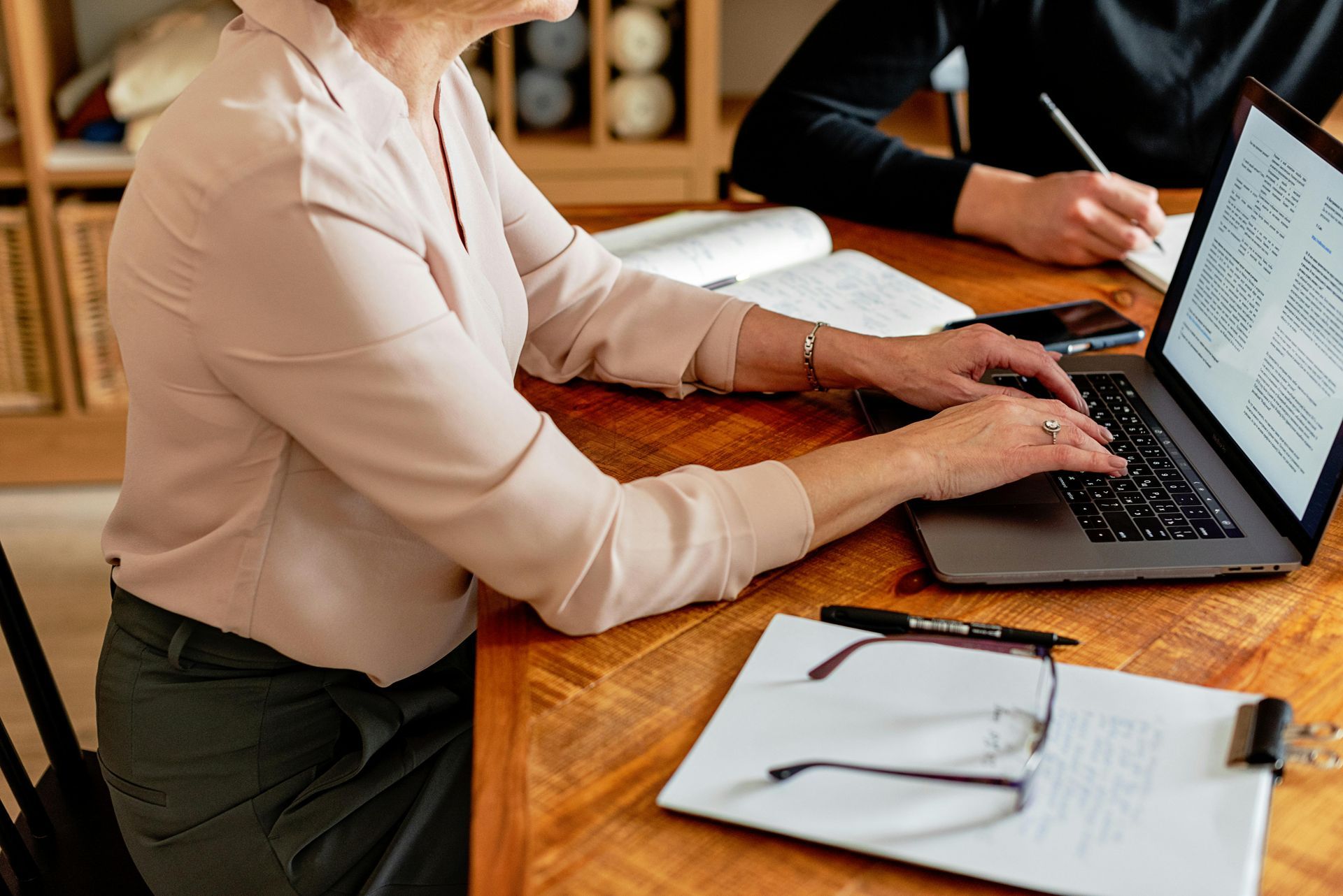 Woman typing on laptop with documents and glasses on a wooden table, another person taking notes.