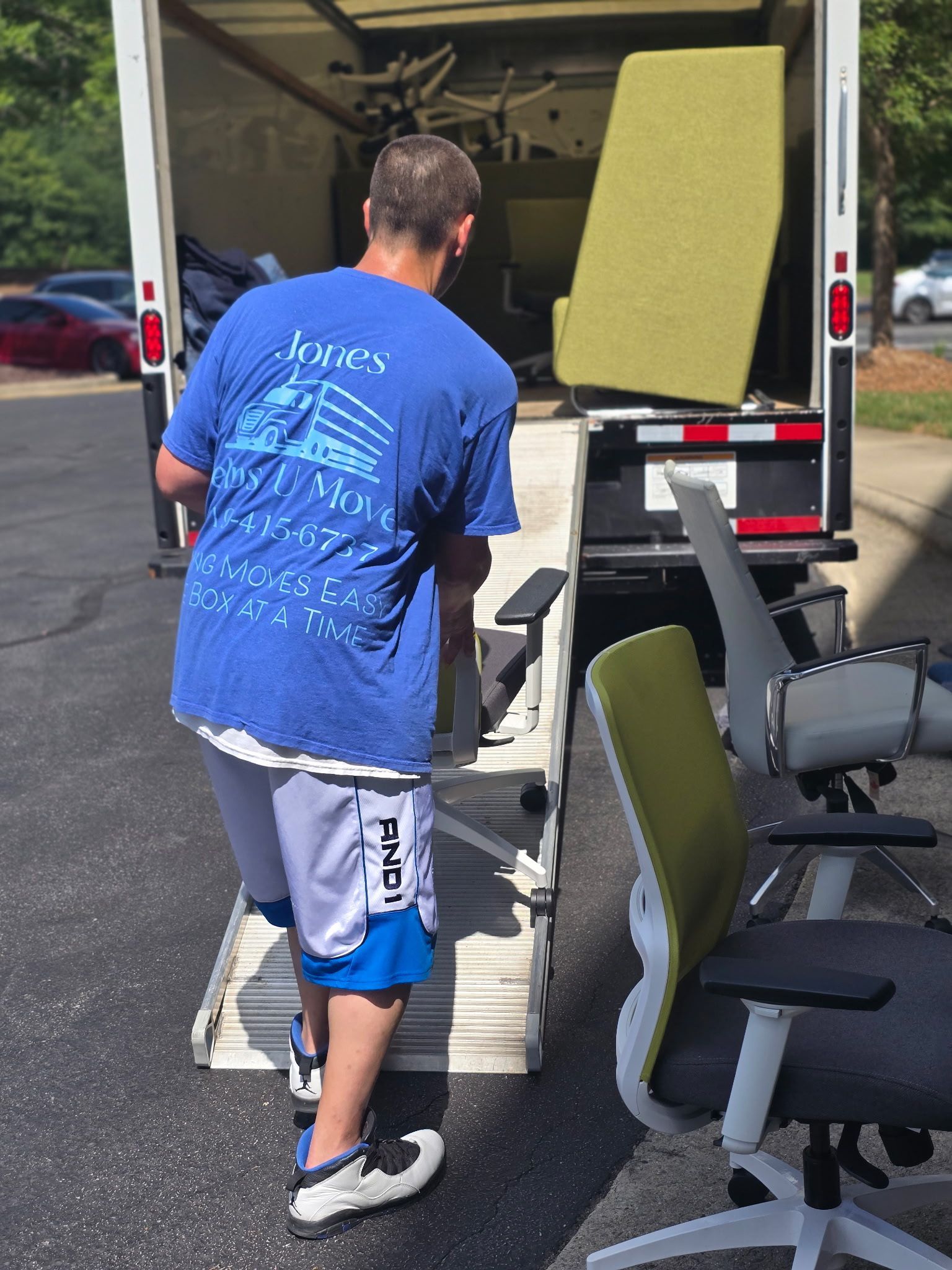 Man loading furniture into a truck via a ramp. A green chair is visible. Sunny, outdoor setting.