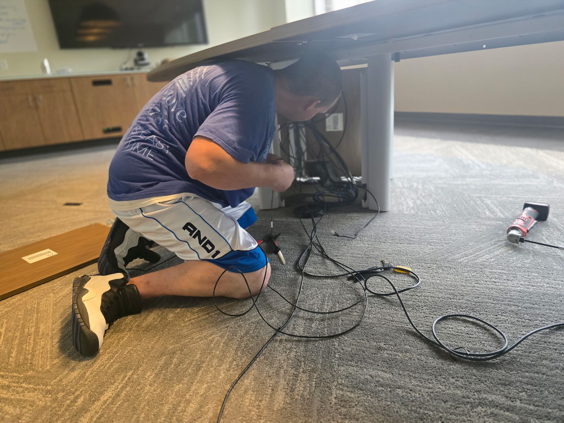 Person kneeling under a desk, working with cables. Gray carpet and tan furniture in an office.