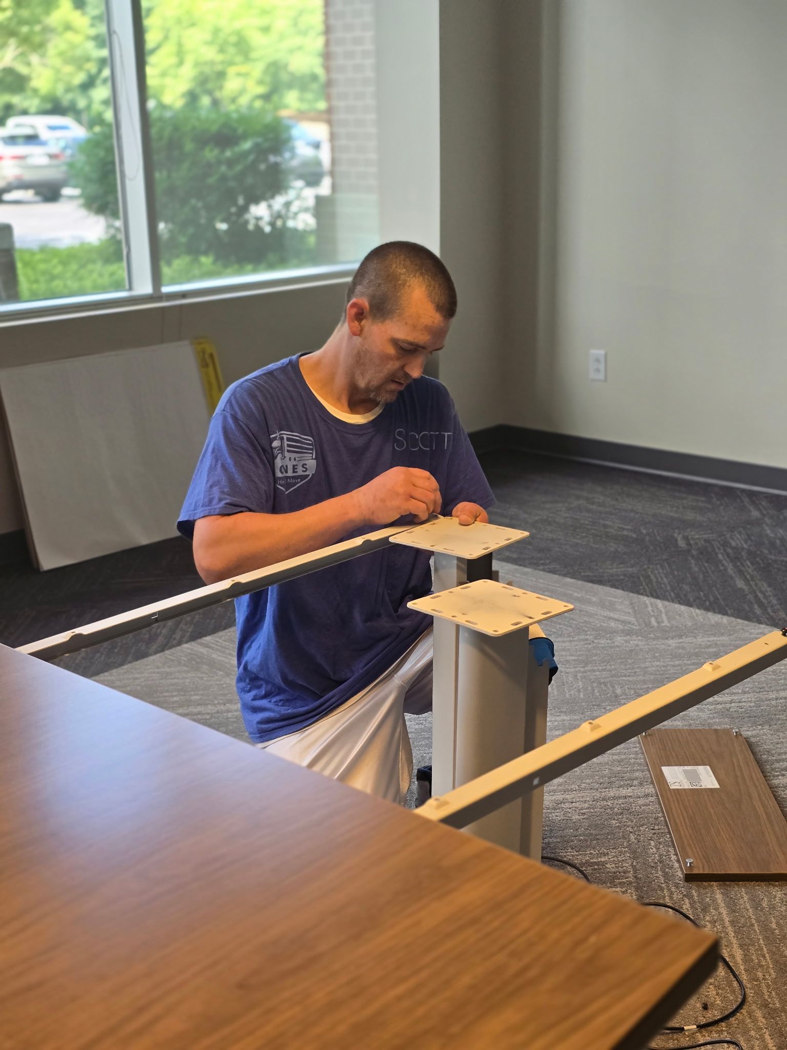 Person kneeling, assembling office furniture, near a window. He is wearing a blue shirt.