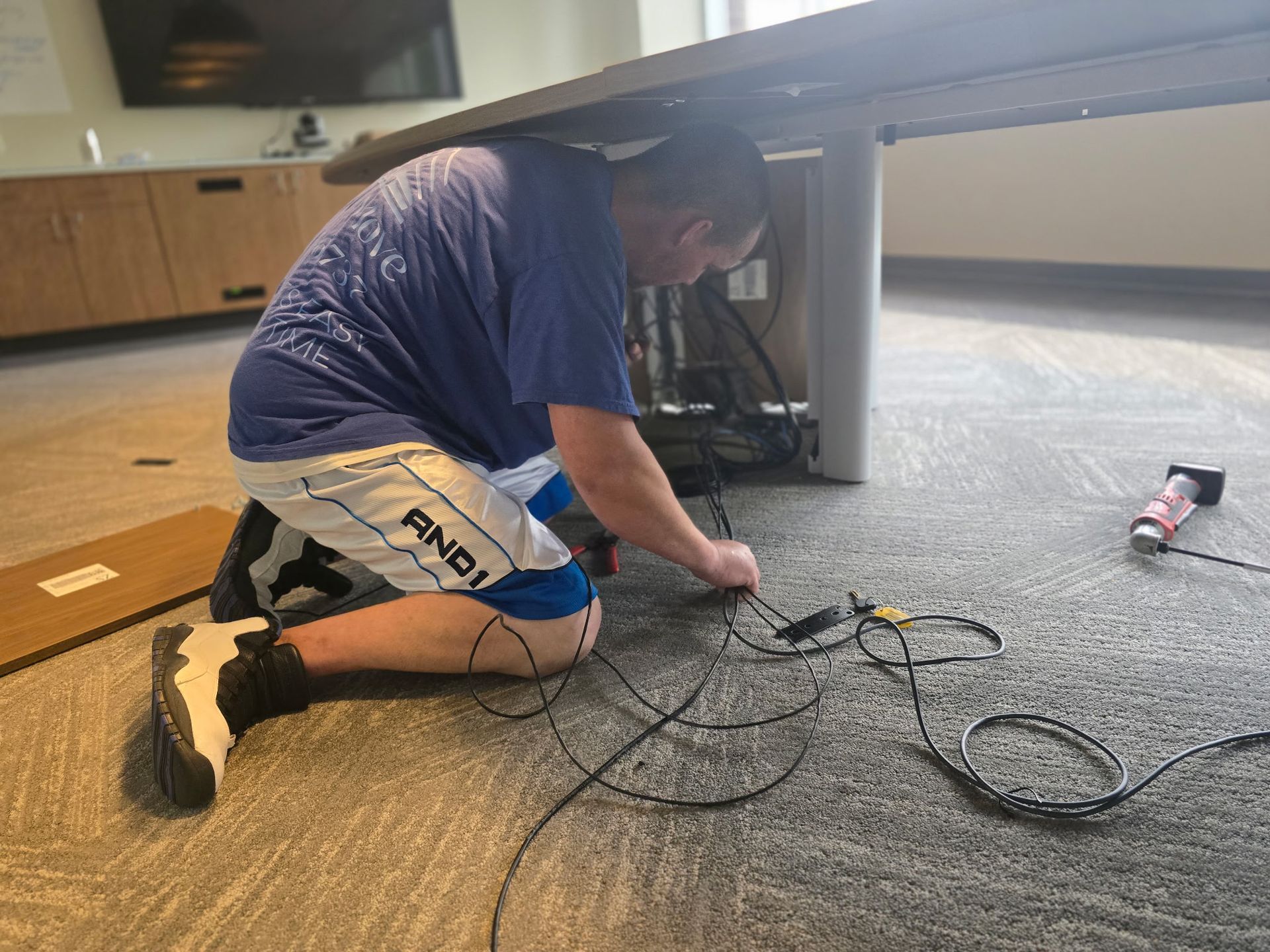 Person kneeling under a table, connecting wires. They wear a blue shirt, shorts, and sneakers on a carpeted floor.