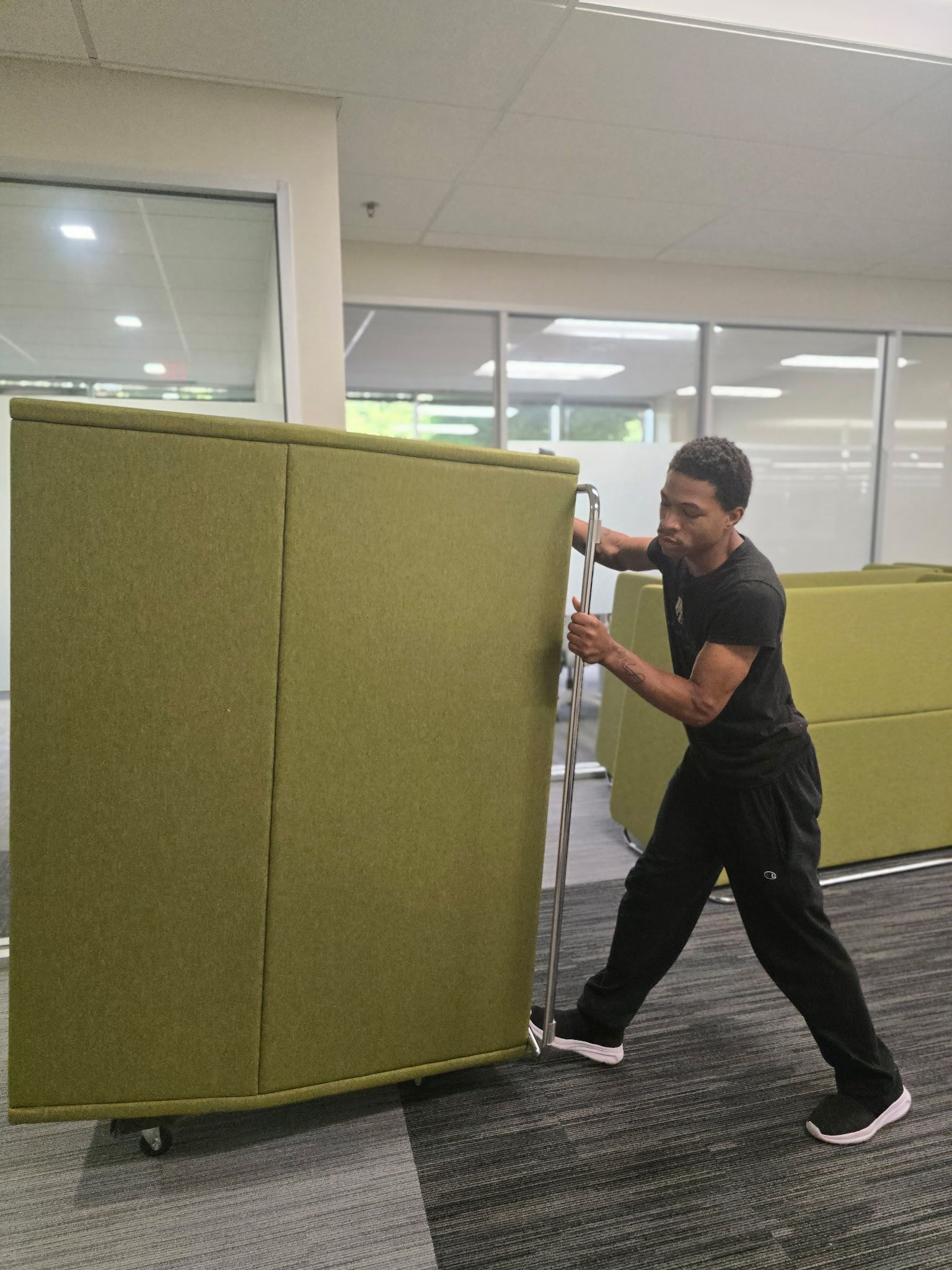 Man pushing a large green upholstered cube on wheels in a modern office space.