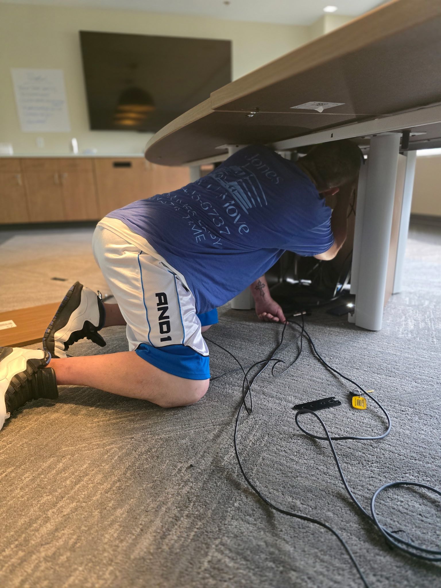 Person kneeling under a table, working on wires. Wearing blue shirt, white shorts, and sneakers. Indoors.