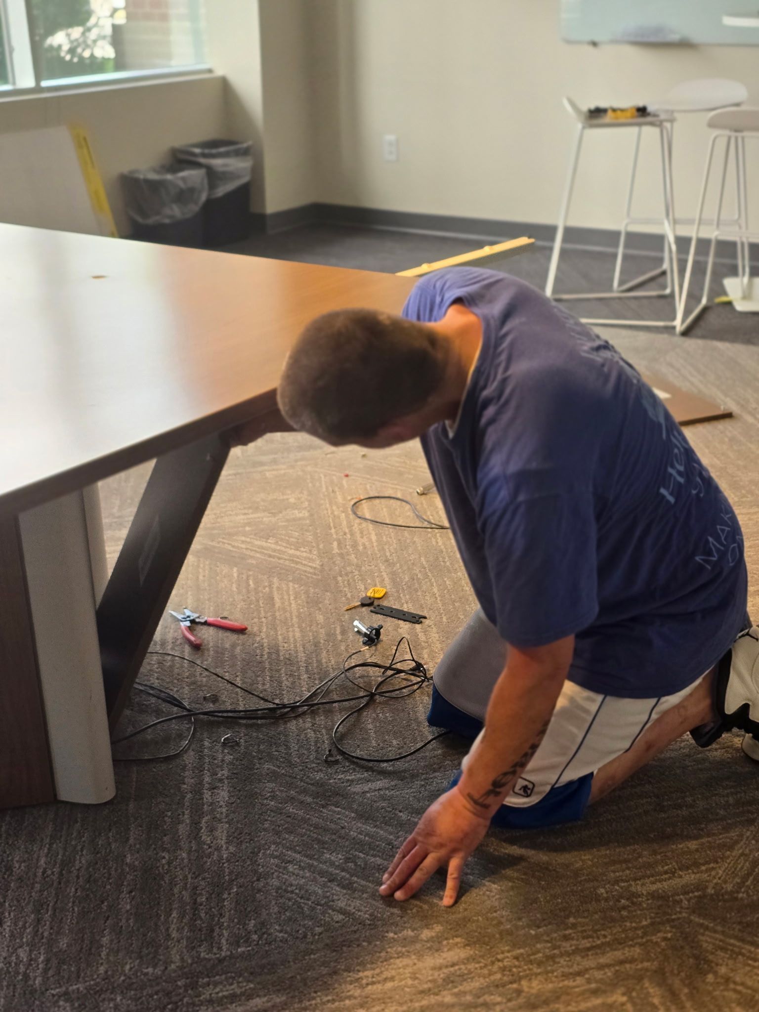 Man kneeling, working under a brown table on a carpeted floor. Cables and tools are nearby.