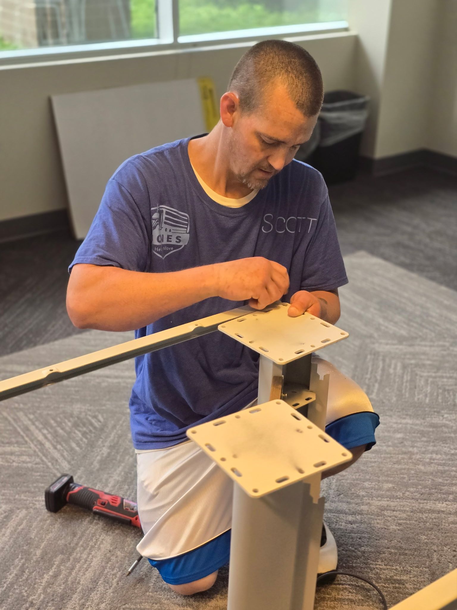 Man assembling furniture, kneeling on a carpeted floor. He wears a blue shirt and white shorts. A window and trash can are in the background.