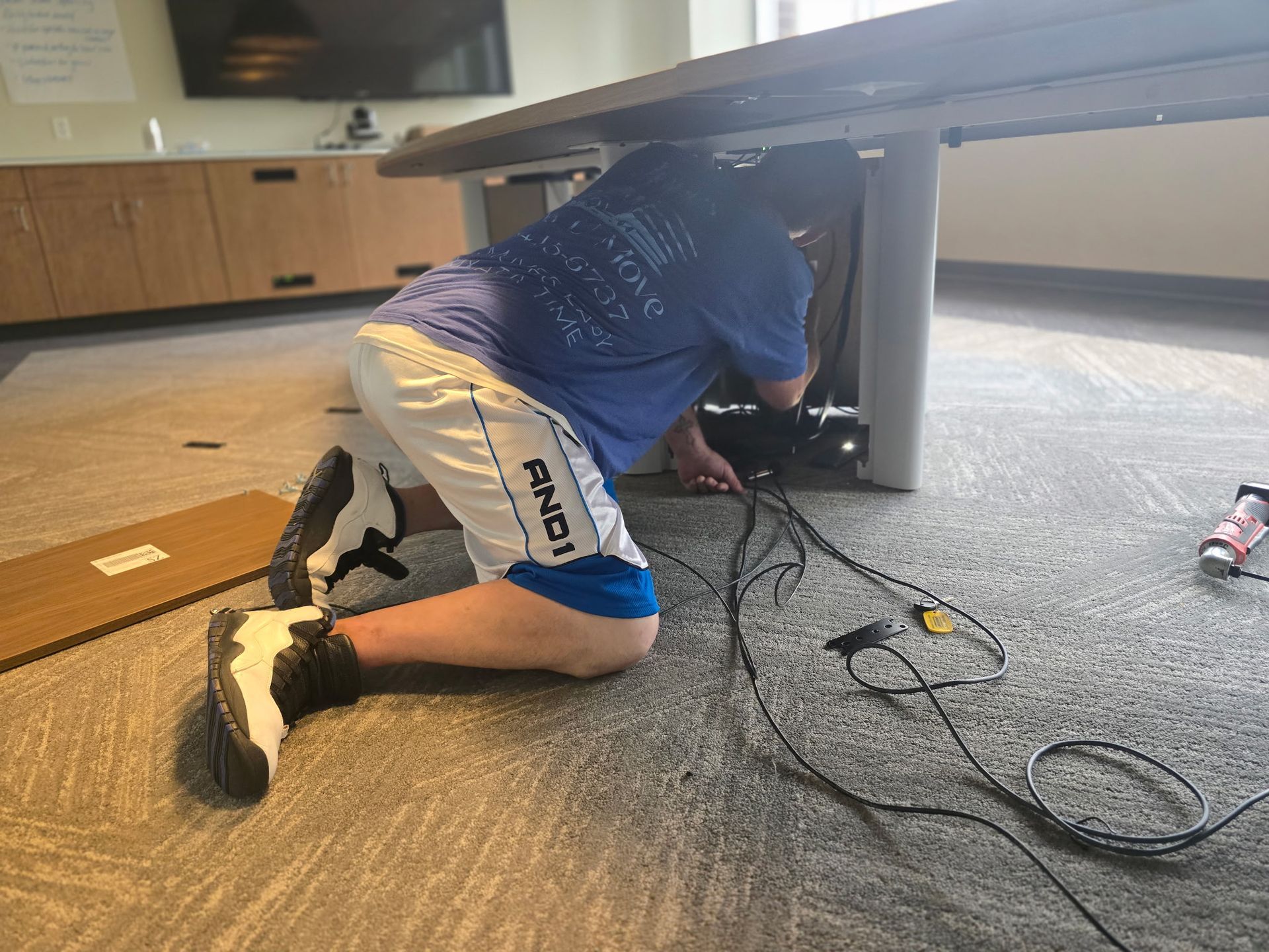 Person kneeling under a table, working on wires. Wears blue shirt, white shorts, and sneakers. Interior office setting.