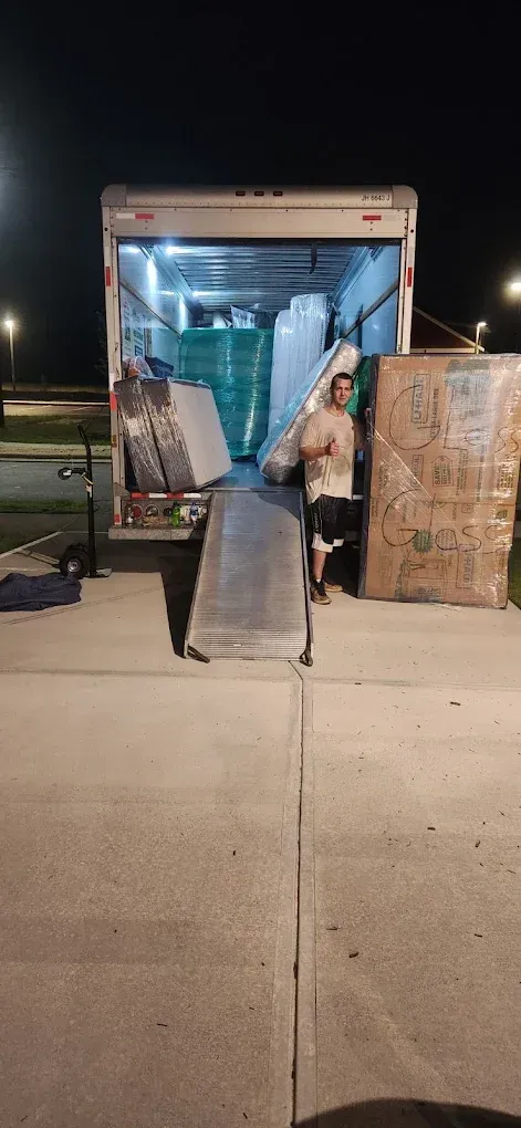 A moving truck with a ramp open at night. A man stands near boxes and the truck's cargo of packed items.