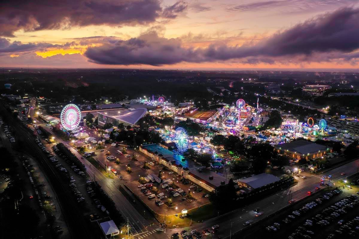 Aerial view of the North Carolina State Fair at dusk, with glowing Ferris wheels and carnival rides under dramatic clouds.