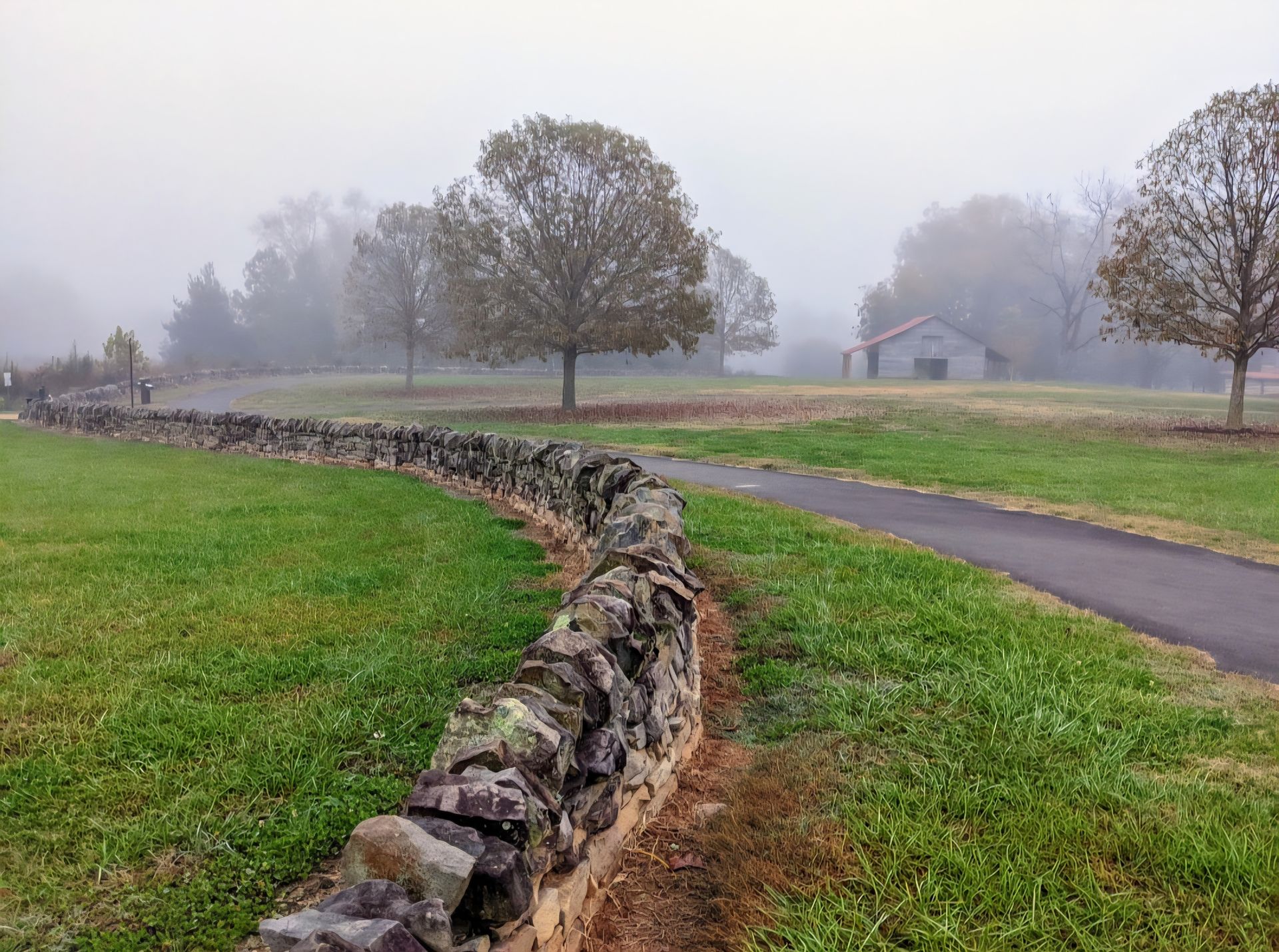A long, rustic stone wall curves through a grassy field next to a paved path at E. Carroll Joyner Park in Wake Forest, NC, on a foggy morning.