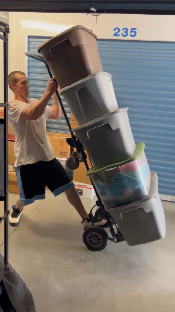 Person pulling a hand truck loaded with stacked storage bins inside a storage unit.