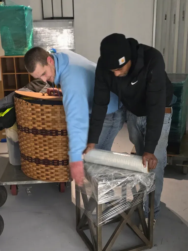Two people wrapping furniture in plastic wrap inside a room.