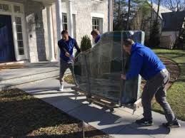 Three movers pushing a large, wrapped object on a dolly up a sidewalk towards a house.