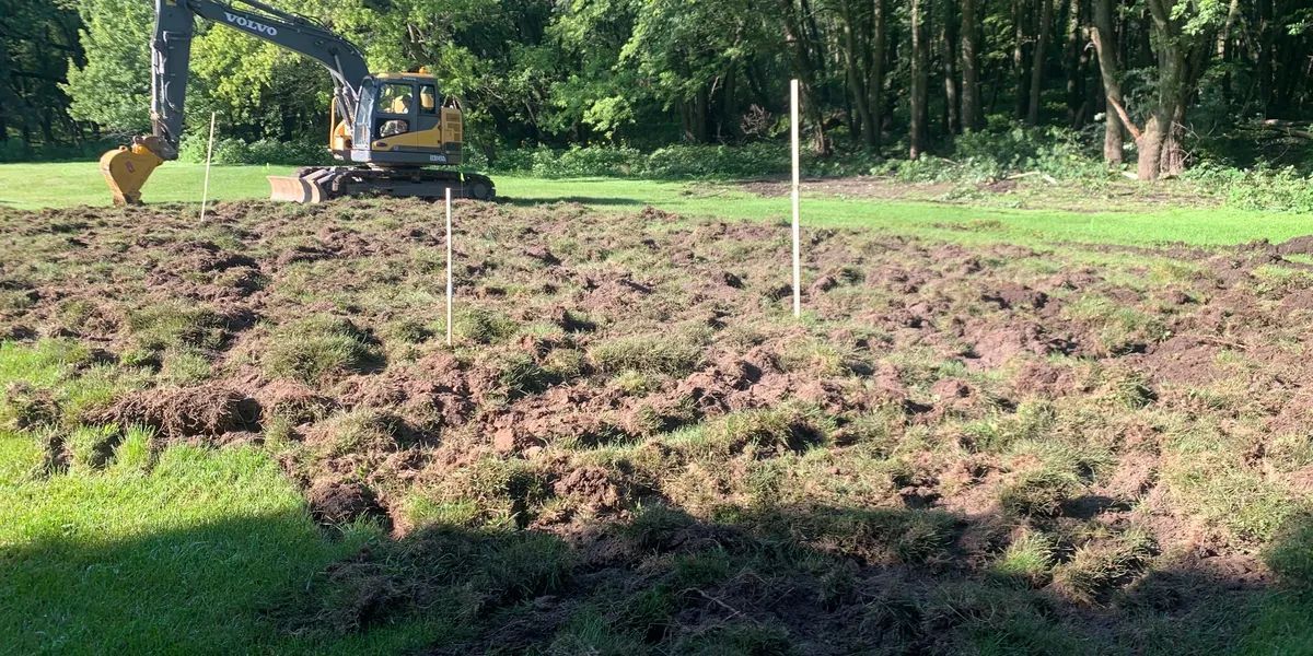 An excavator is tearing up a patch of grass in a field, two white stakes mark the area.