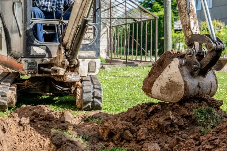 Excavator digging in a yard; dirt mound and tracks visible, greenhouse in the background.