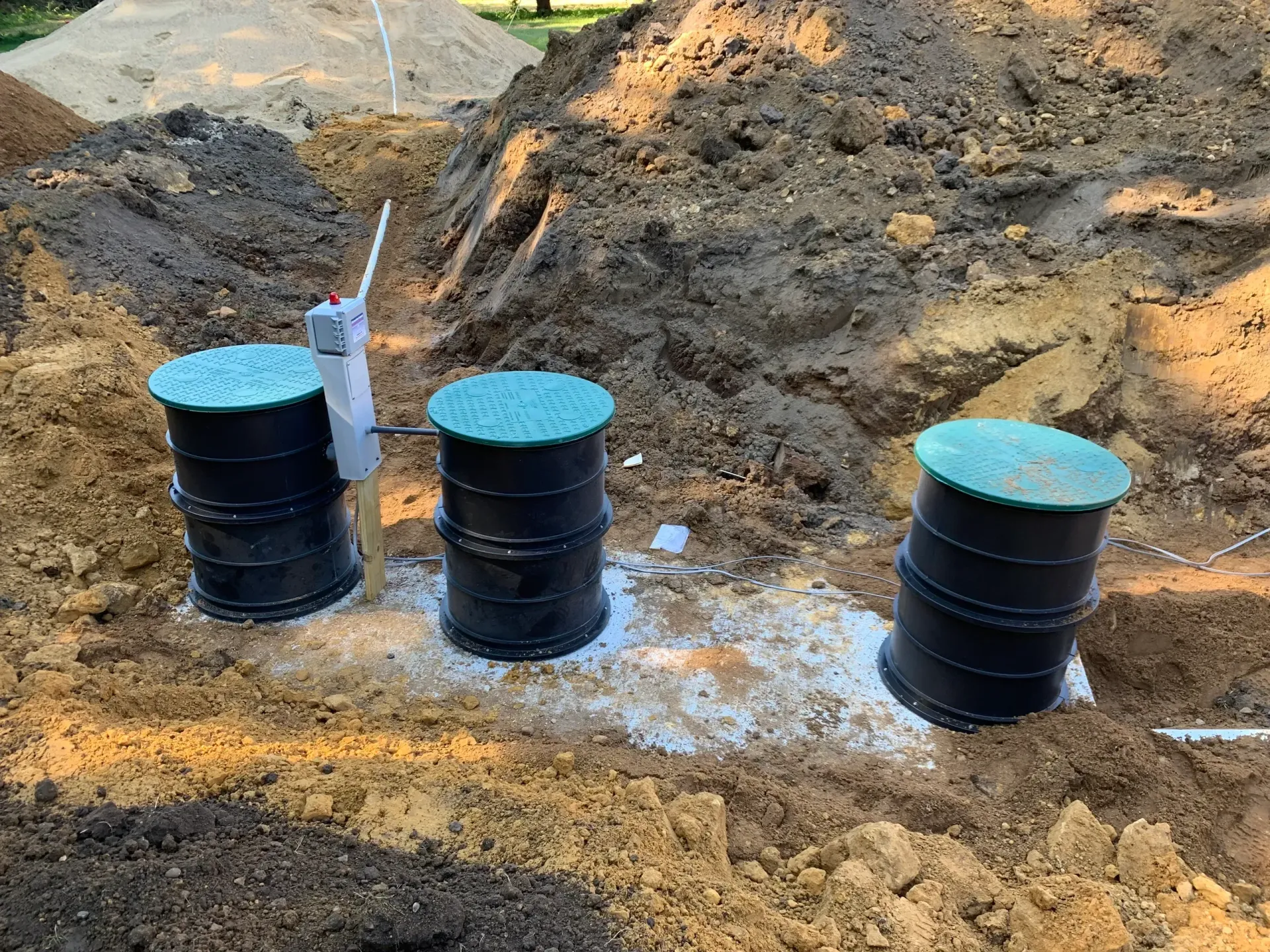 Three black septic tank access points with green lids in an excavated area filled with dirt.