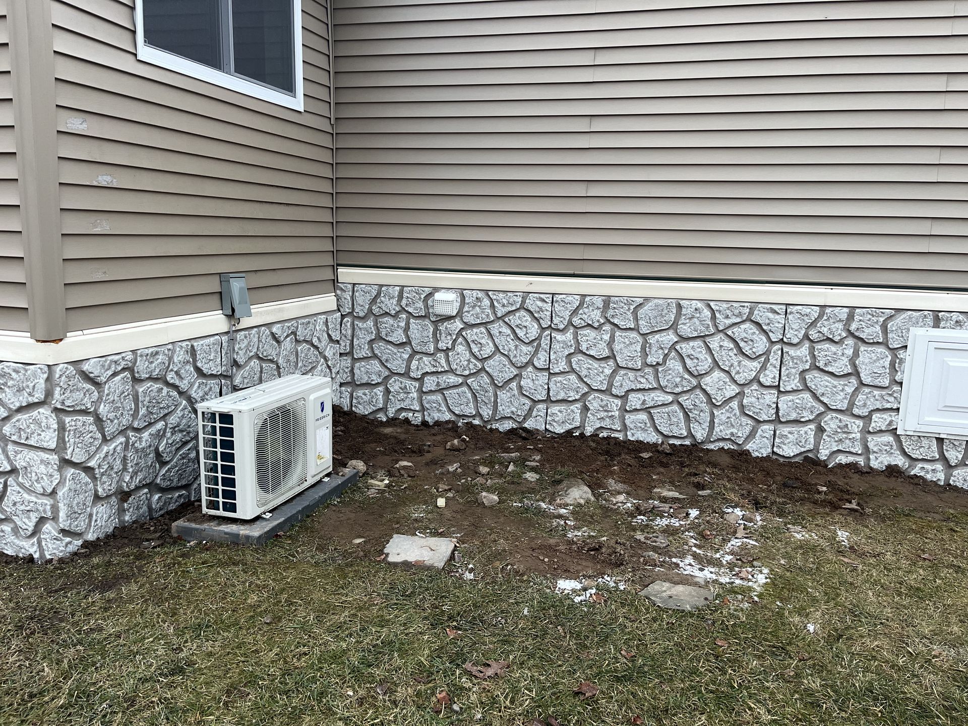 An air conditioner is sitting on the side of a house next to a stone wall.