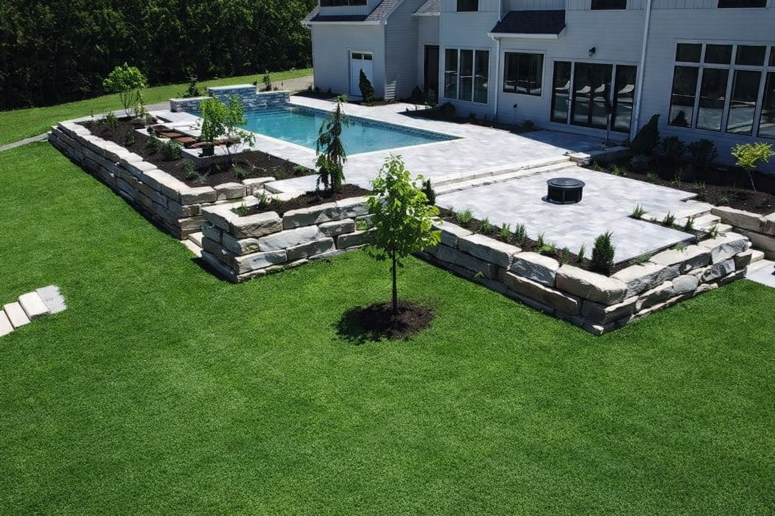 Backyard patio with stone retaining walls, white paving, and a swimming pool beside a house.