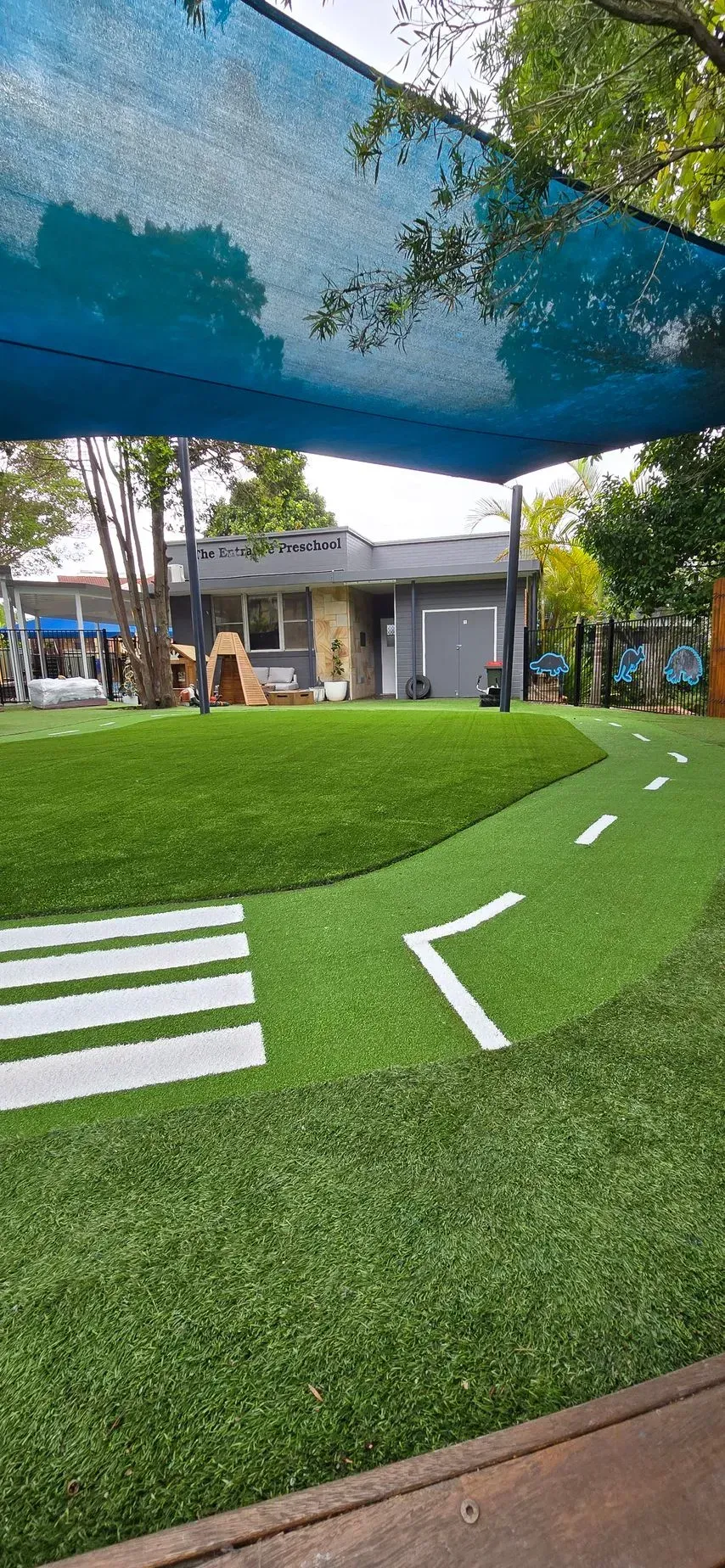 Playground with a grassy hill and a road painted on it. A shaded area with a building in the background.