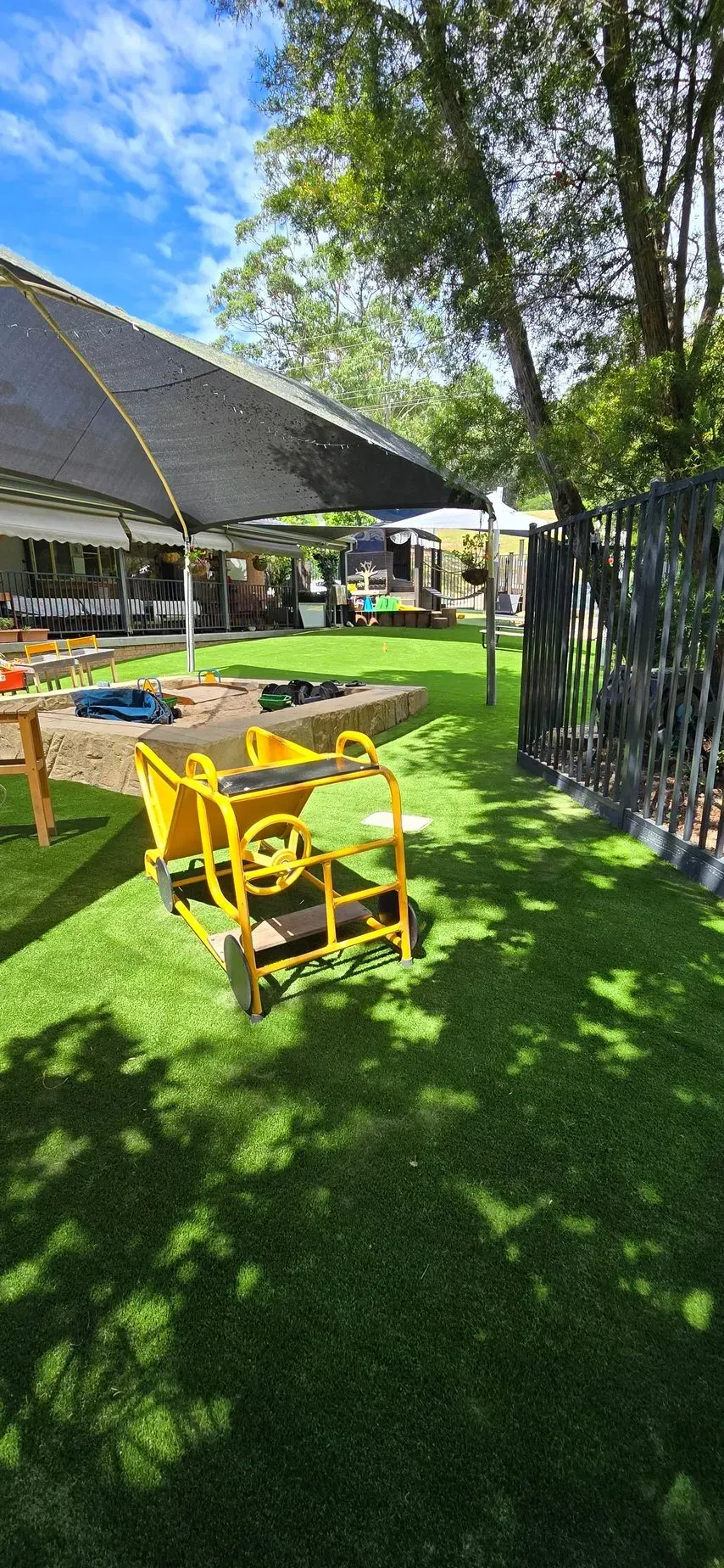 Yellow cart on green grass in a playground with a black shade and trees.