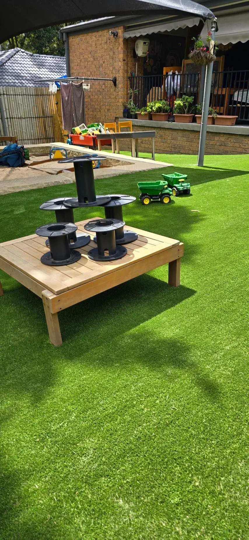 Outdoor play area with artificial turf, wooden table with cable spools, and toy tractors.
