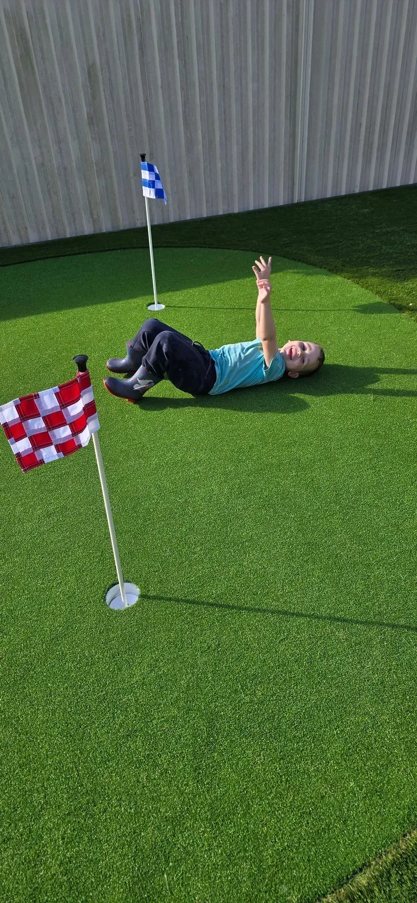 Child on green turf with arms raised, near golf flags. Wooden fence in background.