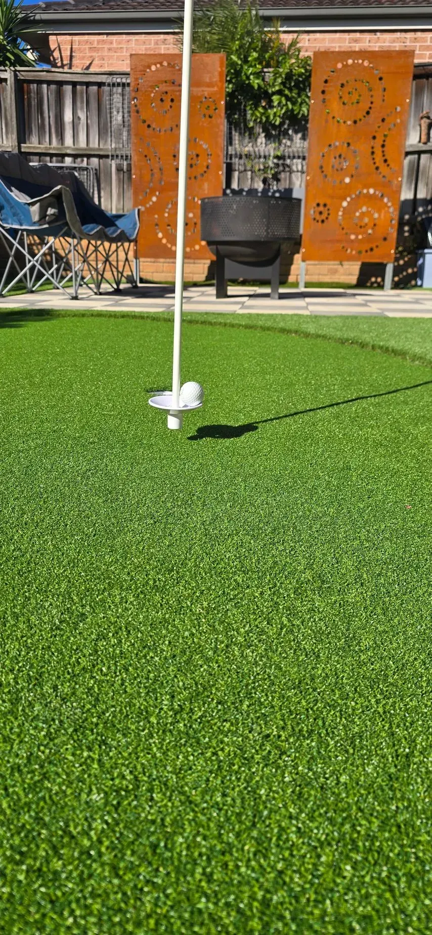 A golf hole with flag on artificial green lawn. Decorative metal screens and a fire pit are in the background.